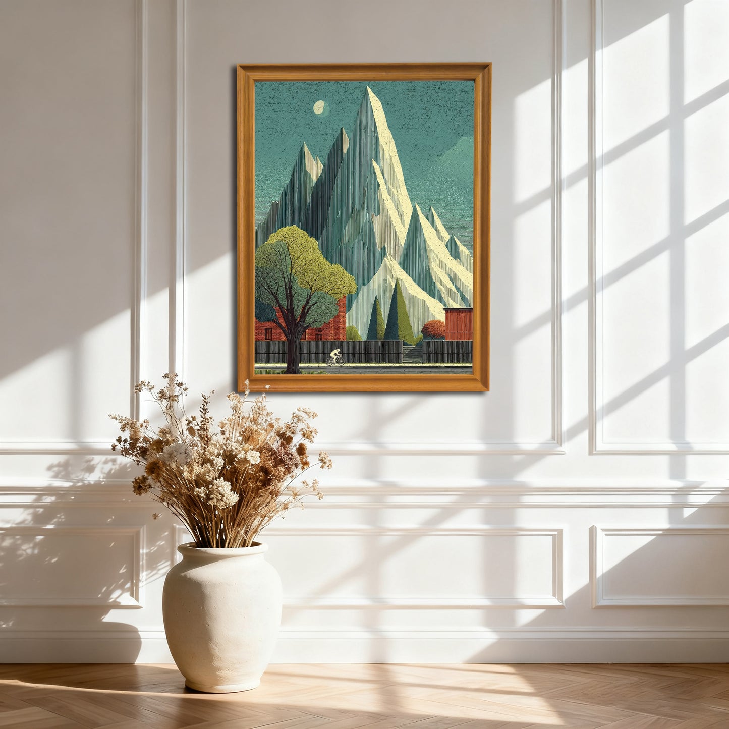 Natural wood-framed art print featuring sharp white mountains and a cyclist passing a large tree. The print is hanging on a white paneled wall with dramatic sunlight and shadows.