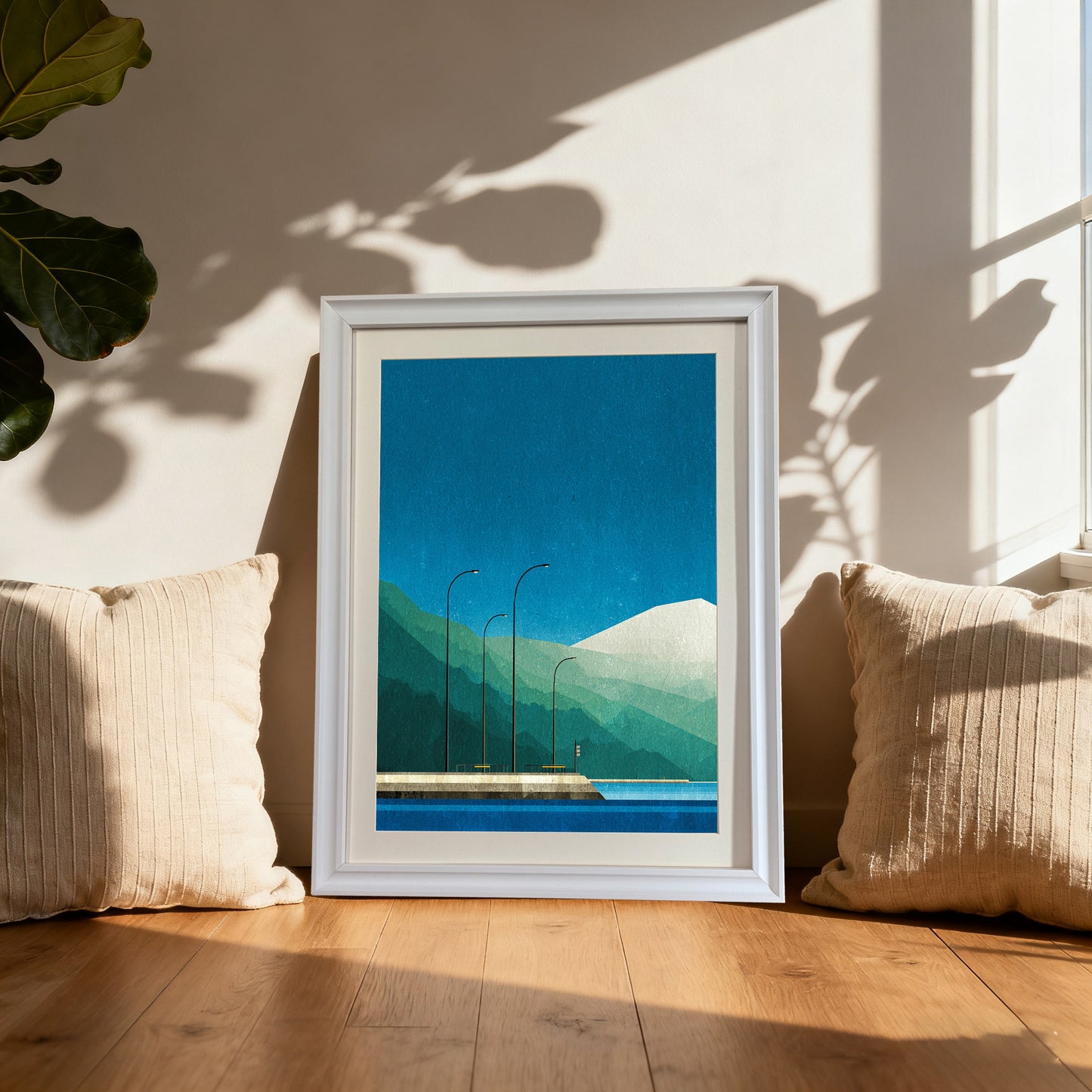 Geometric art print of a blue and green mountain landscape, framed in white, leaning on a wooden floor between two cream-colored pillows by a sunlit window.