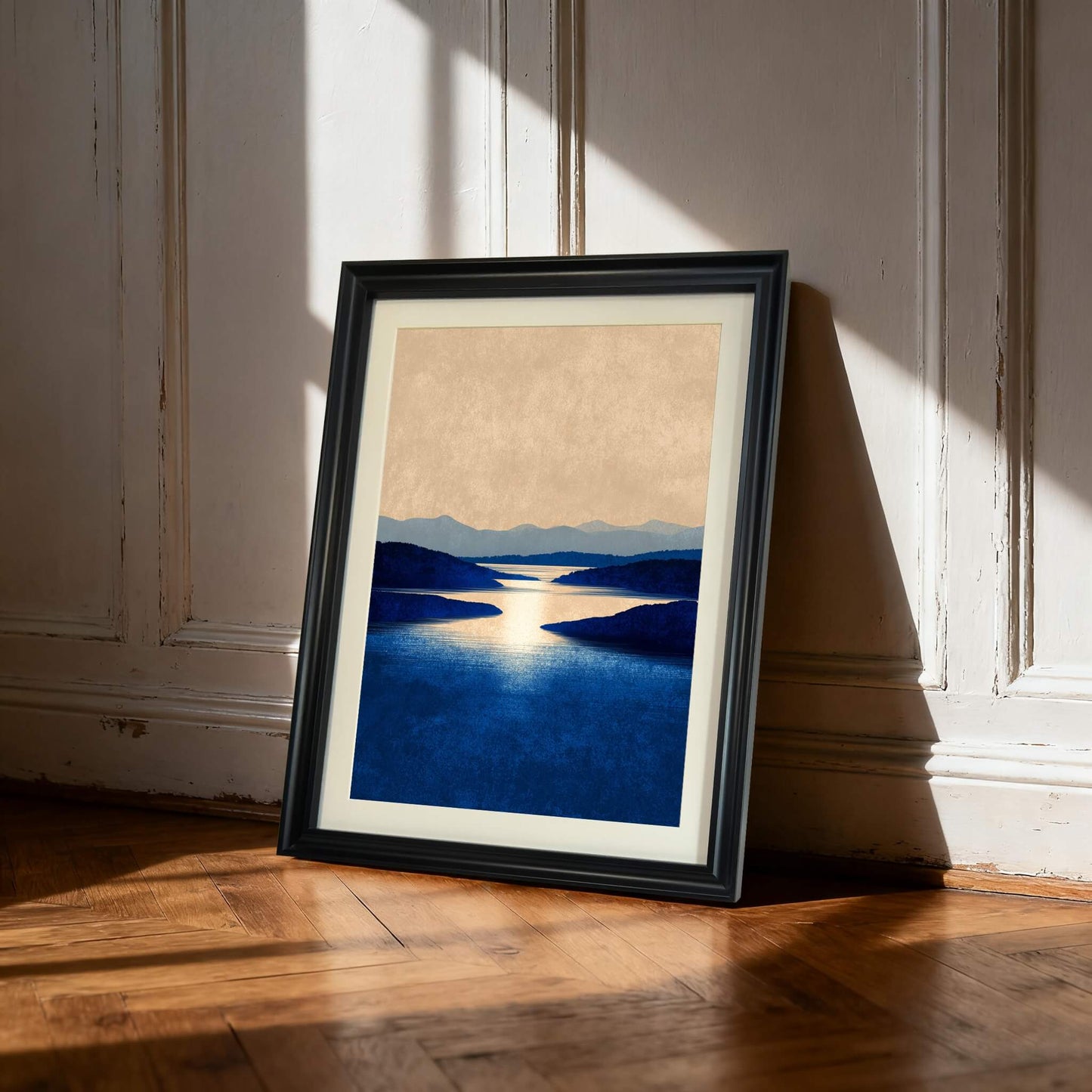 Art print of a dark blue lake with sun reflection and layered hills, displayed in a black frame, leaning against a white paneled wall on a wood floor.