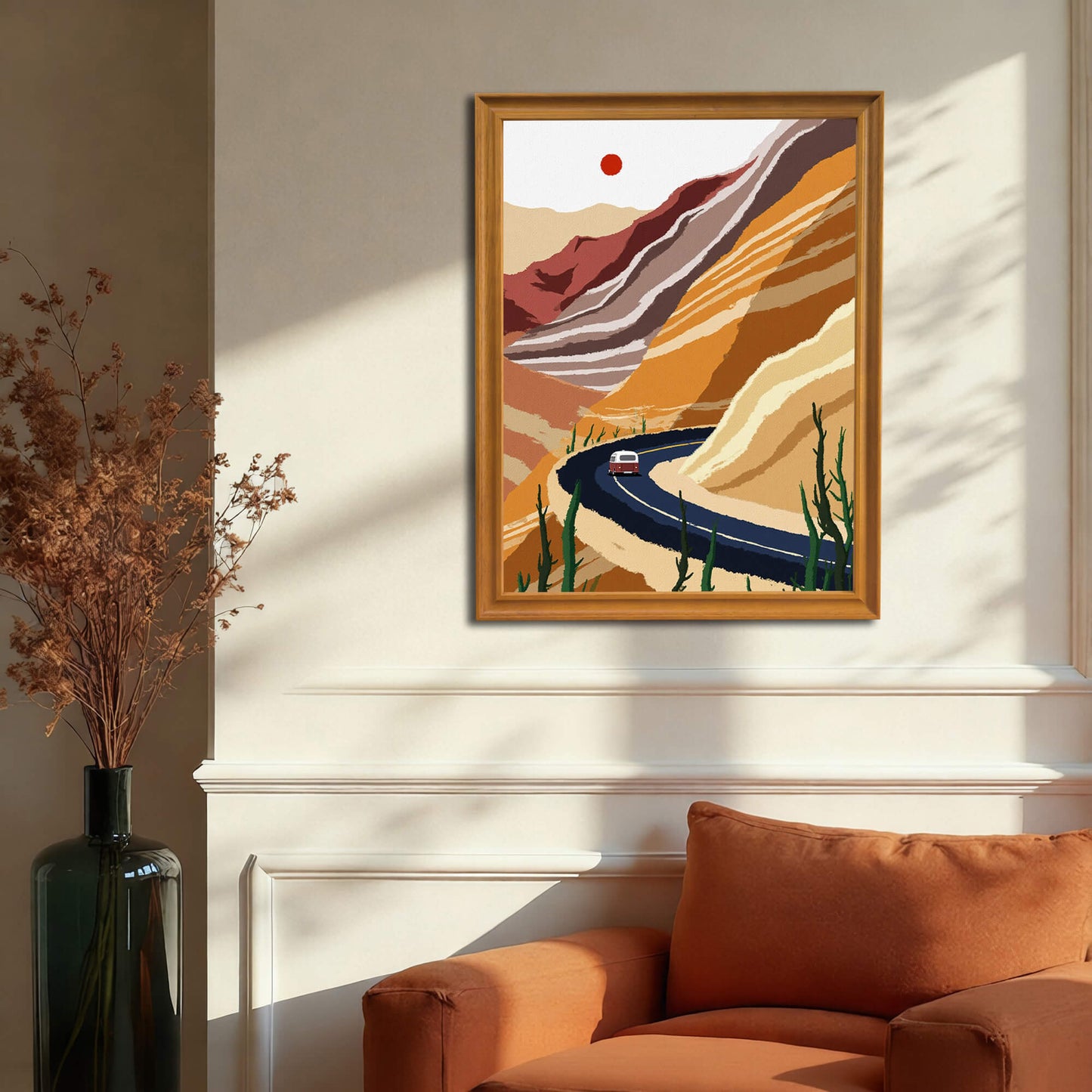 The desert road art print, framed in warm wood, hanging on a cream wainscoting wall above a rust-orange armchair. A dark vase with dried flora stands next to the chair.