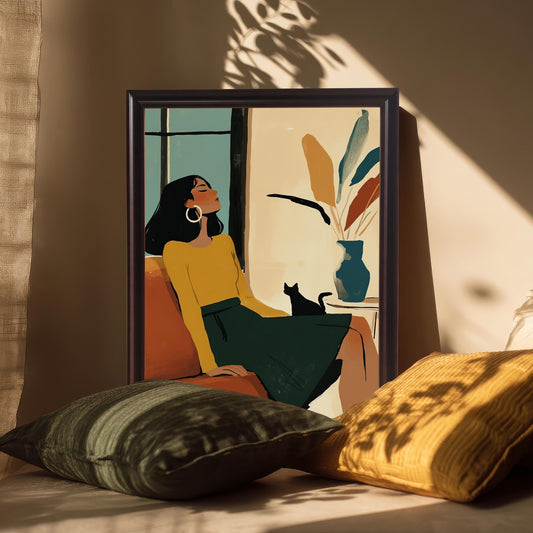 A framed art print of a woman and cat, with a black wood frame, propped on a bench with throw pillows. Sunlight casts shadows from a window across the art.