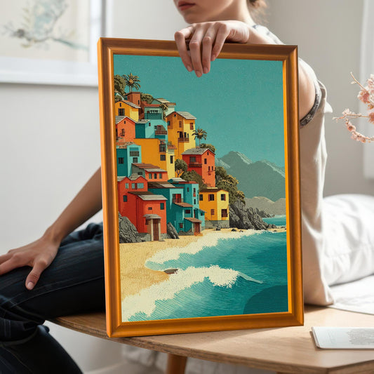 A person sitting on a table holds up a framed art print. The print shows a collection of colorful houses on a sandy beach next to a turquoise ocean.