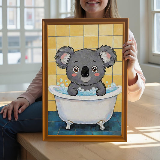 A person holds a framed art print of a koala in a bathtub. The frame is orange with no mount. The person is sitting on a wooden surface in front of a window.