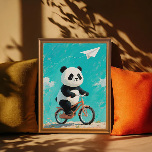 A framed print of a cute panda riding a bicycle, with a blue sky and a paper airplane. The frame is a warm, light brown. The image is a close-up, with orange and olive-green pillows surrounding the frame.