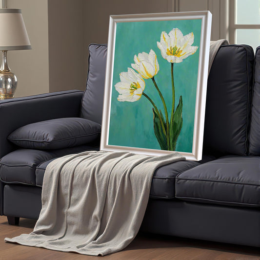 A framed art print of three white tulips with green leaves on a teal background, leaning on a dark gray couch. The print has a white frame and a white mat. A gray blanket is draped on the couch.