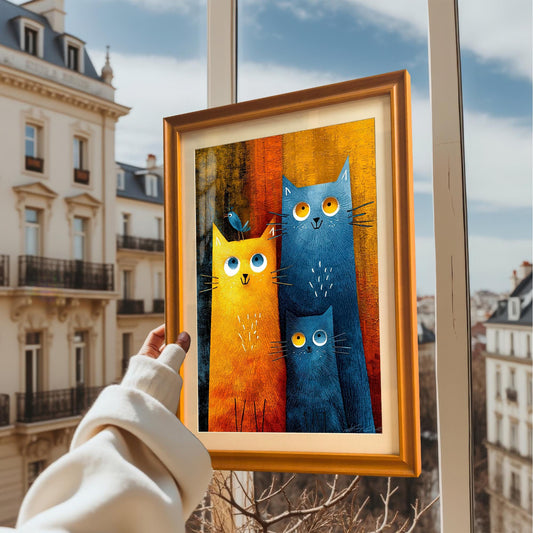 A hand in a white sleeve holds up the framed cat family print against a background of a sunny city view with buildings and a bright blue sky. The frame is a bright orange-gold.