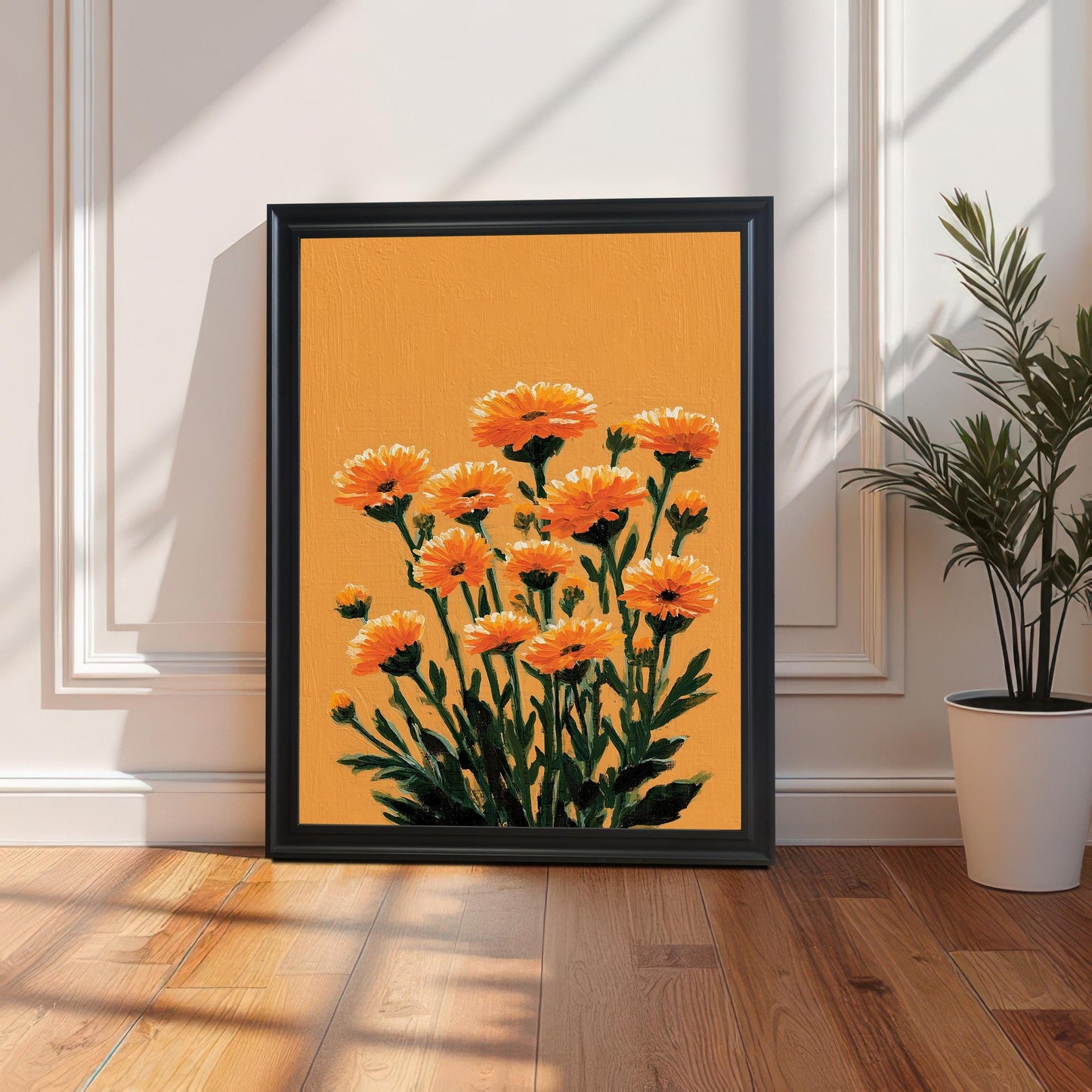 A portrait-oriented orange floral art print in a Black frame leaning on a light wood floor against a white paneled wainscoting wall, next to a potted houseplant.