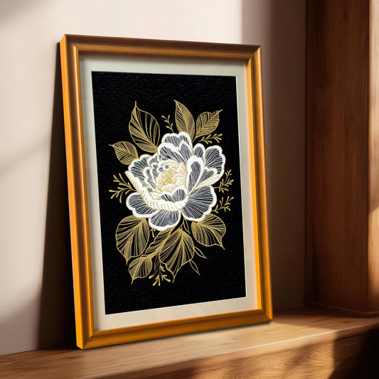 A framed black and gold floral print with a gold-colored frame, leaning on a light-colored wooden windowsill with sunlight streaming in from the right.