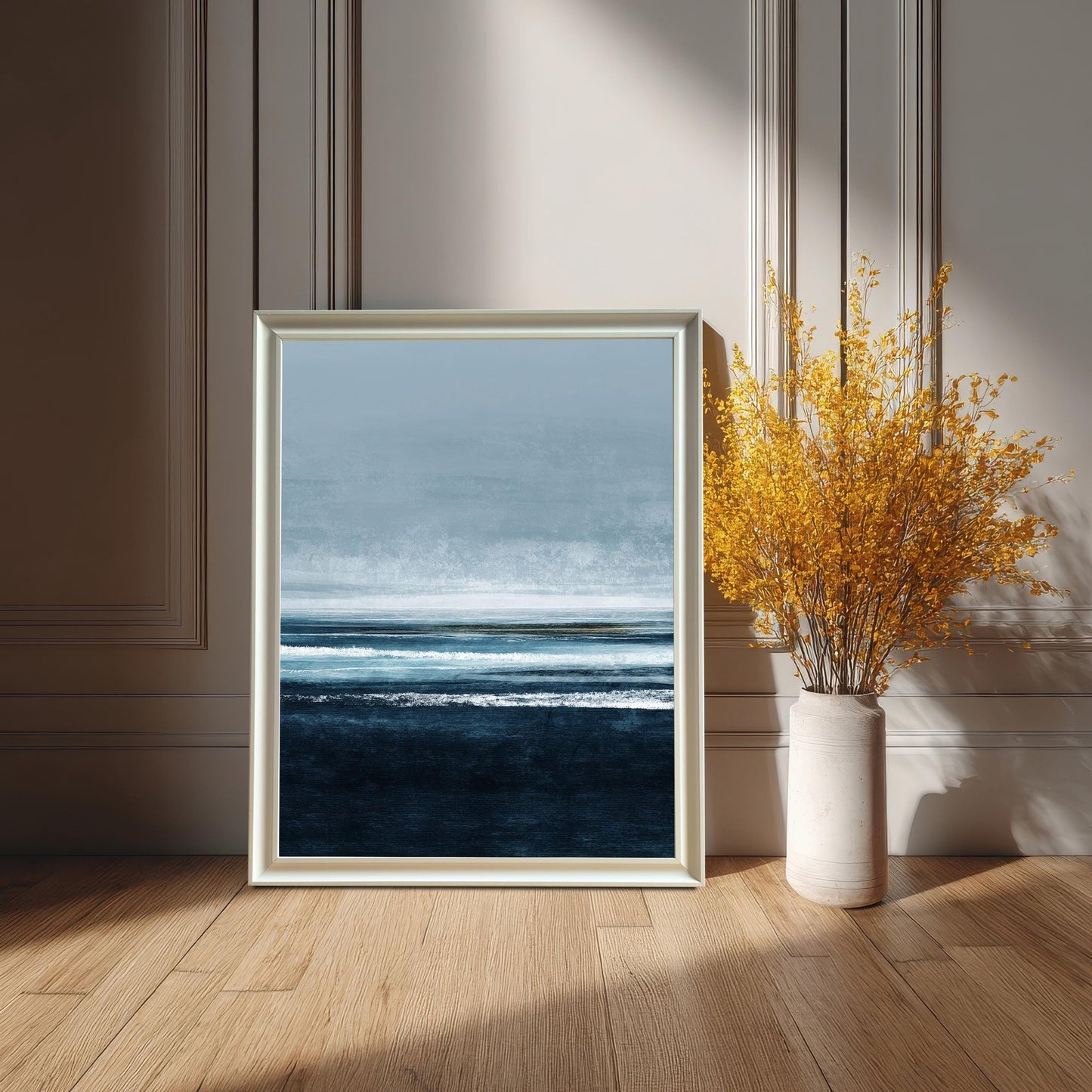 Abstract modern seascape art print featuring deep navy blue waves and light blue sky, in a White frame leaning on a wooden floor against a white paneled wall. A tall white vase with bright yellow dried flowers is beside it.