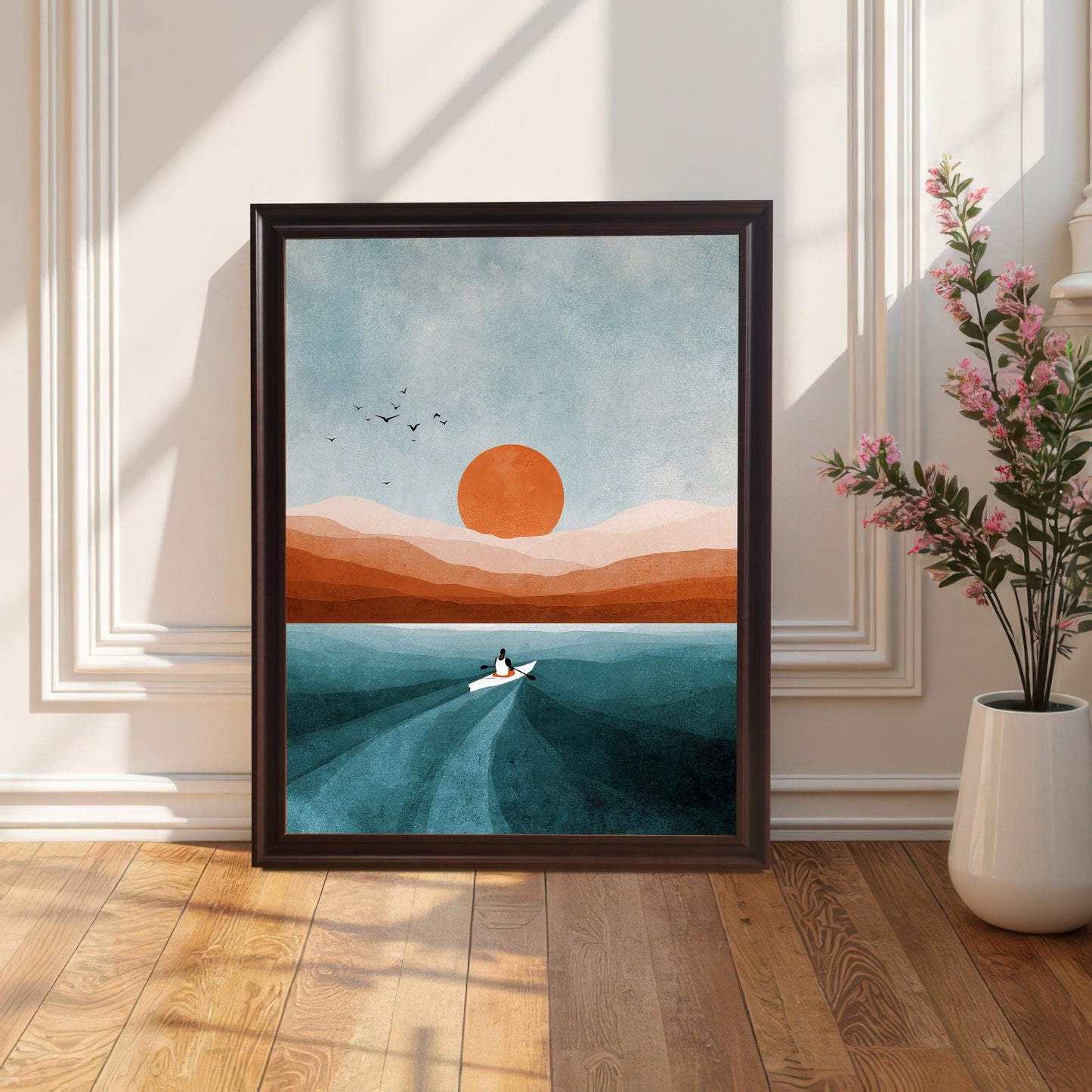 Framed art print in a Coffee (dark brown) wood frame (no mount), leaning on a sunlit wooden floor against a white paneled wall. A white vase with tall pink flowers is on the right. Shows the print in a classic/transitional decor setting.