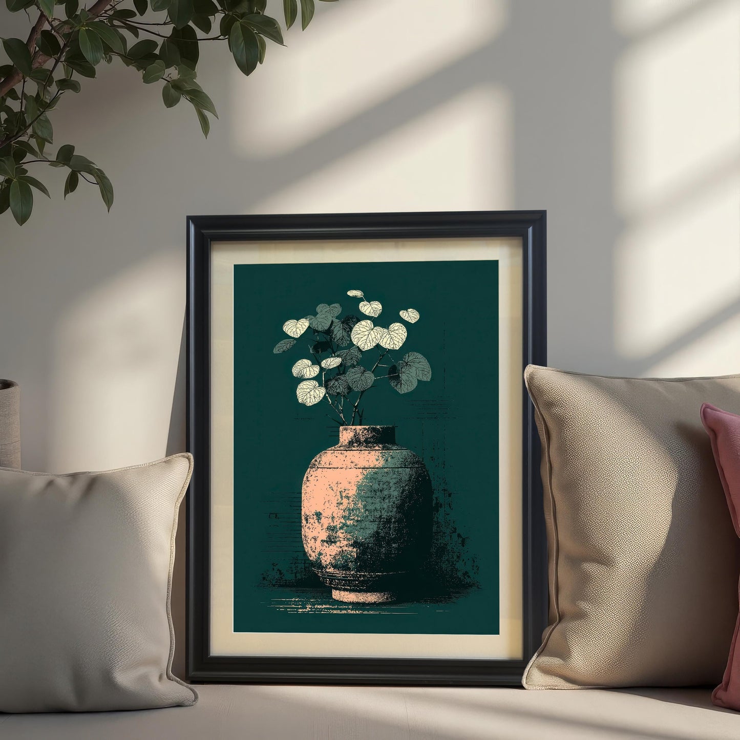A framed art print of a distressed vase with pale leaves on a dark teal background, resting on a cushioned surface between two beige throw pillows. The frame is Black with a cream-colored mat. A plant hangs from the top-left corner, and sunlight casts strong shadows across the scene.