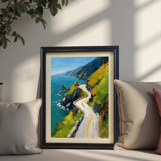 A framed print of a vibrant oil painting of a winding coastal road, resting on a cushioned surface between two beige throw pillows. The frame is Black with a cream-colored mat. A plant hangs from the top-left corner, and sunlight casts strong shadows across the scene.