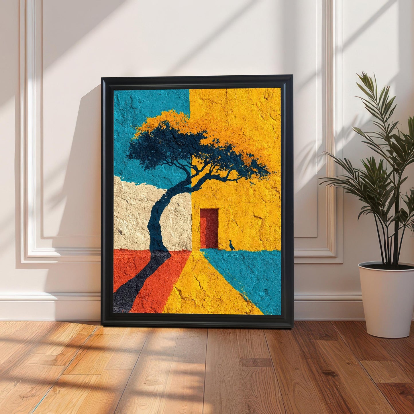 A framed art print in a black frame, leaning on a wooden floor against a white paneled wall (wainscoting). The art is brightly colored and features a tree silhouette, a red door, and blocks of yellow, blue, and red texture. Sunlight casts long, dramatic shadows across the floor. A potted green plant sits to the right.