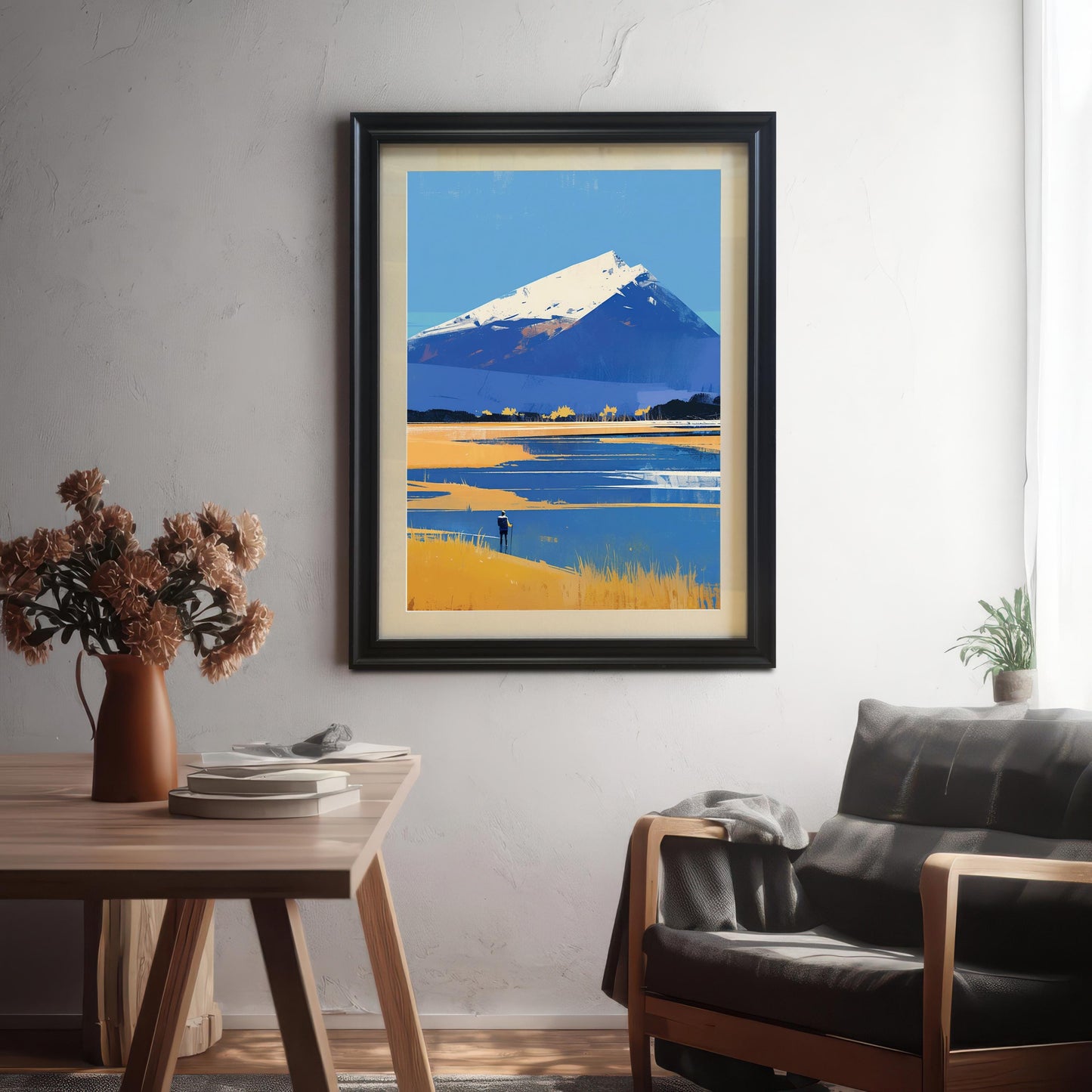 A large mountain landscape art print in a sleek black frame with an off-white mount, hanging on a light gray textured wall. The scene is set in a modern living room featuring a dark leather chair with a gray throw blanket and a wooden table with a vase of dried flowers in the foreground.