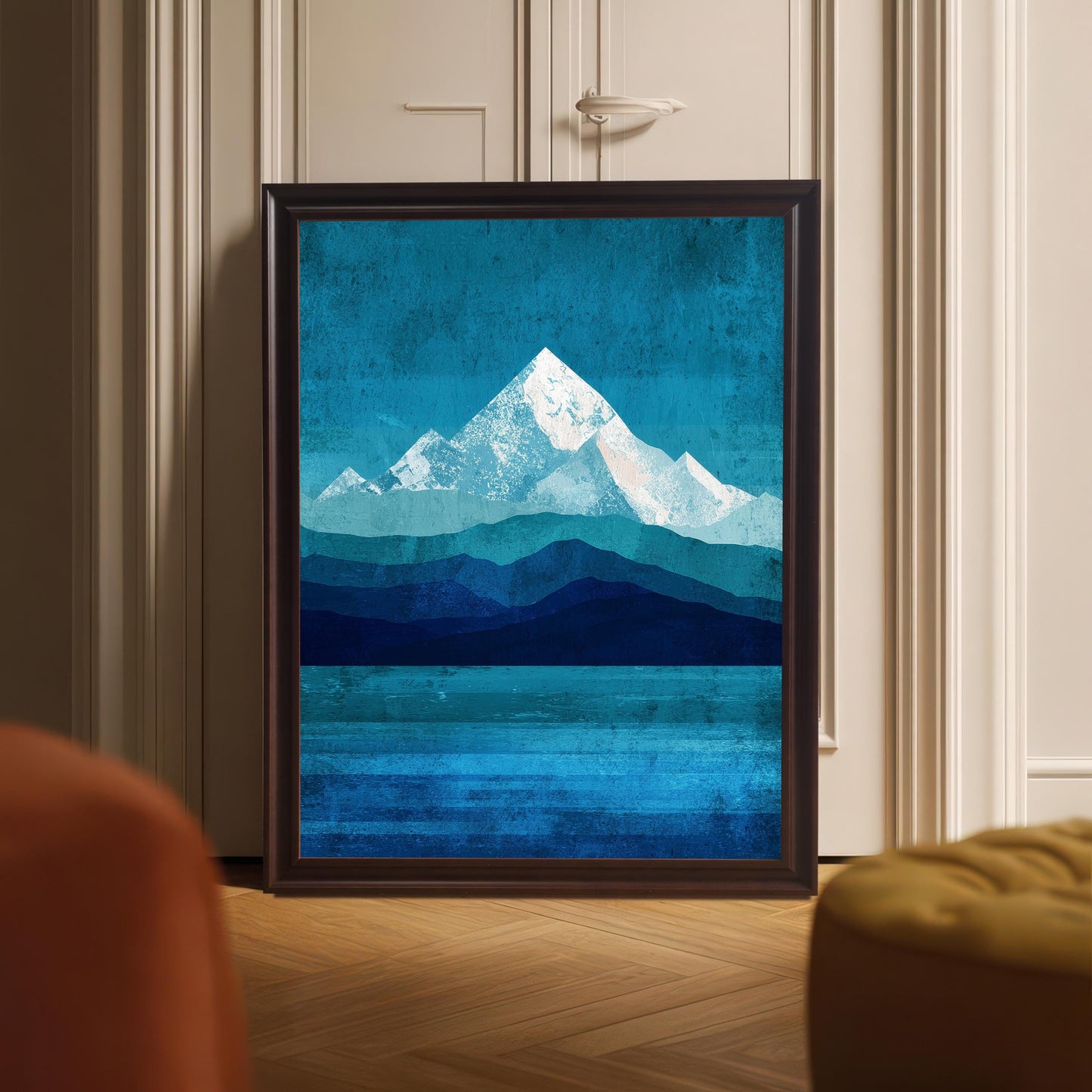 A full-bleed mountain landscape print in a deep "Coffee" brown wood frame, leaning directly on a light wood floor against a neutral paneled door. The mountain peaks are depicted in shades of white and teal over a layered blue-toned sea.