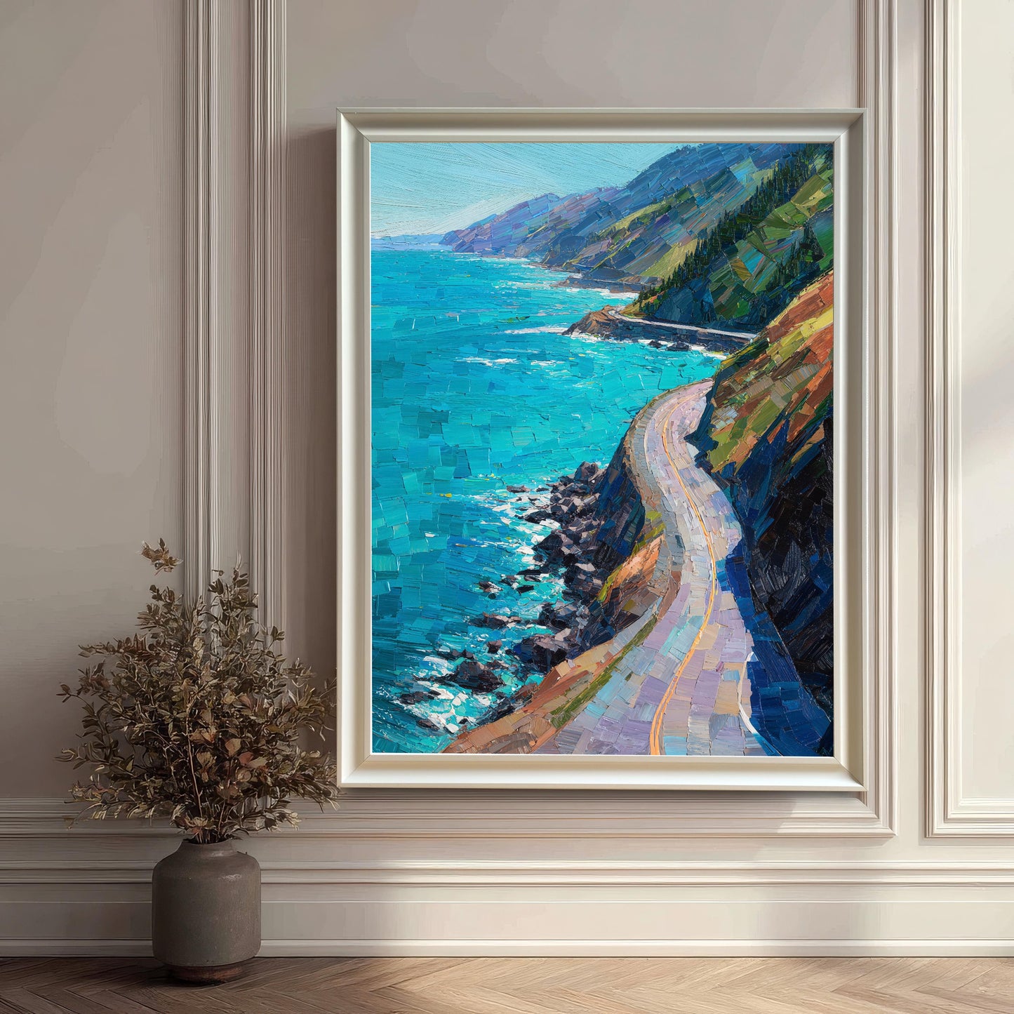 A white wood framed vertical art piece showcasing a vibrant blue ocean and coastal highway, mounted on a white wainscoted wall next to a dark gray vase with decorative dried branches.