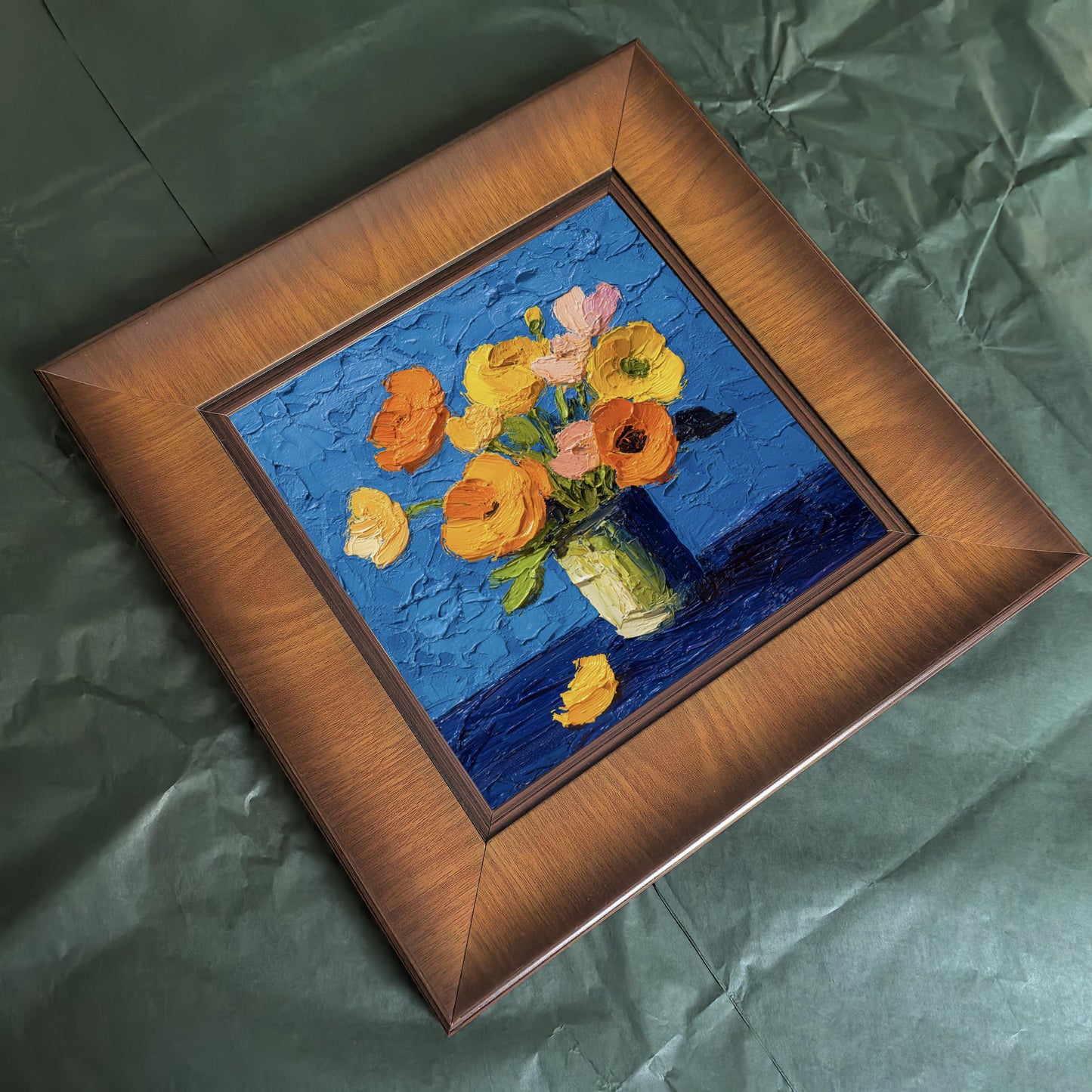 A square wood-framed impasto oil painting of yellow and orange flowers in a vase against a vibrant blue textured background, resting on green paper.