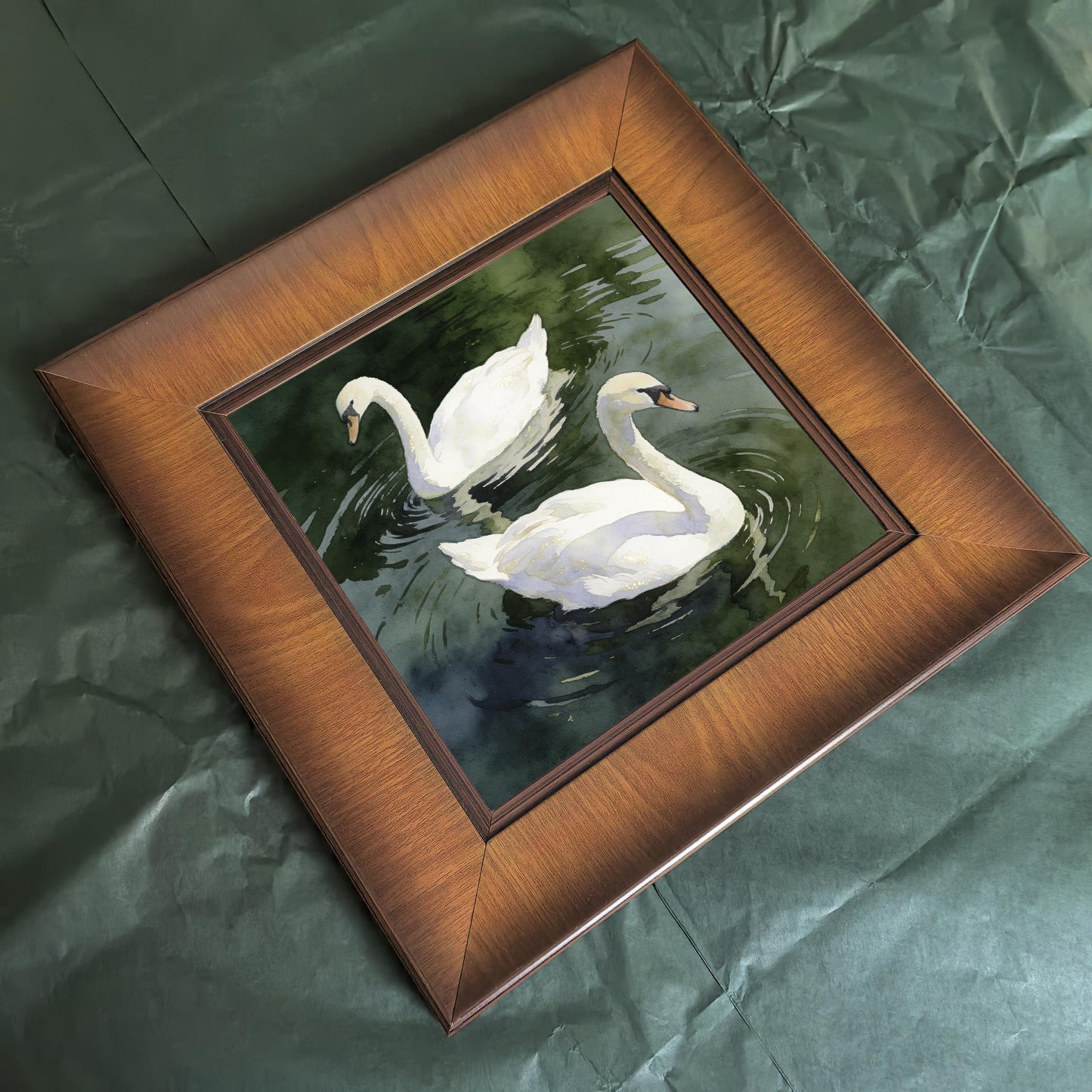 Angled flat lay view of the square wooden frame featuring the swan watercolor print, resting on a dark green crumpled paper surface.