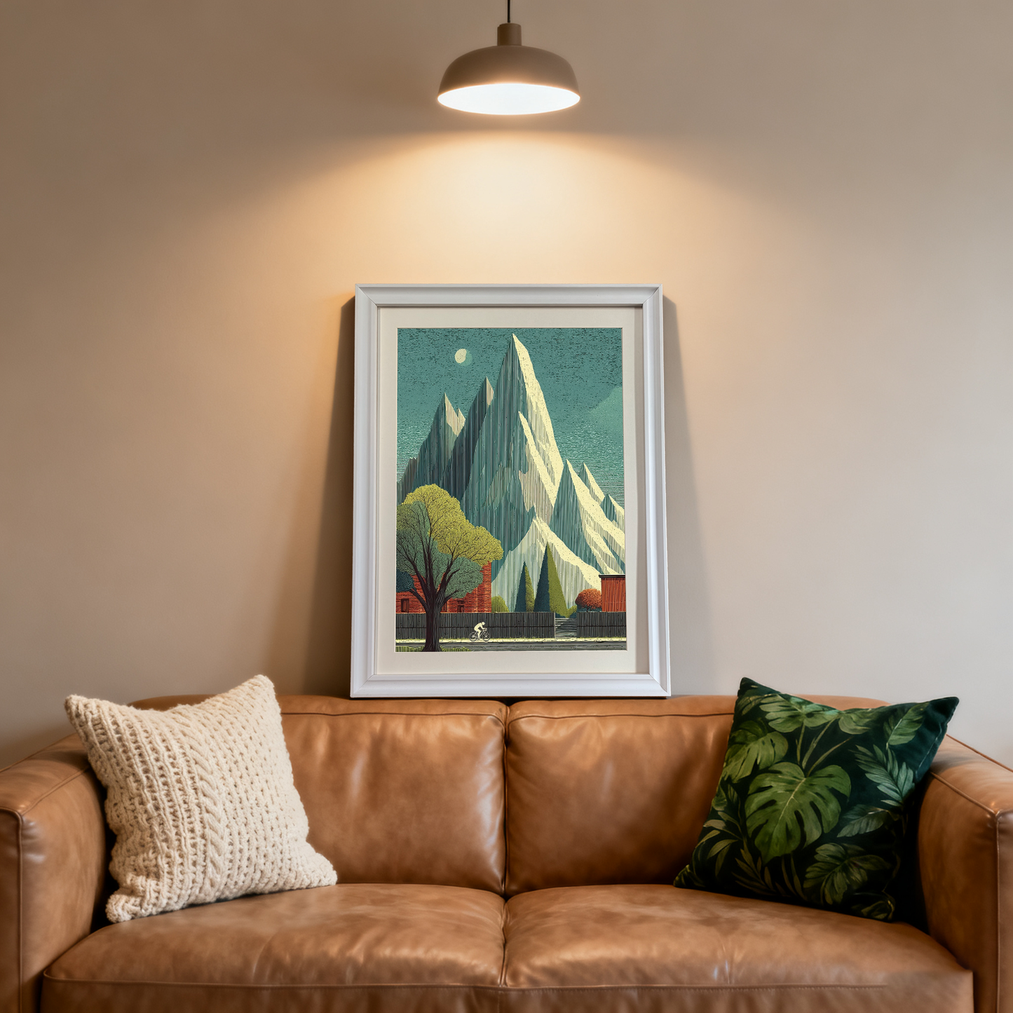 White-framed art print of a striking mountain landscape with a lone cyclist, a large tree, and red buildings. It rests on a brown leather couch in a modern room, illuminated by a spotlight.