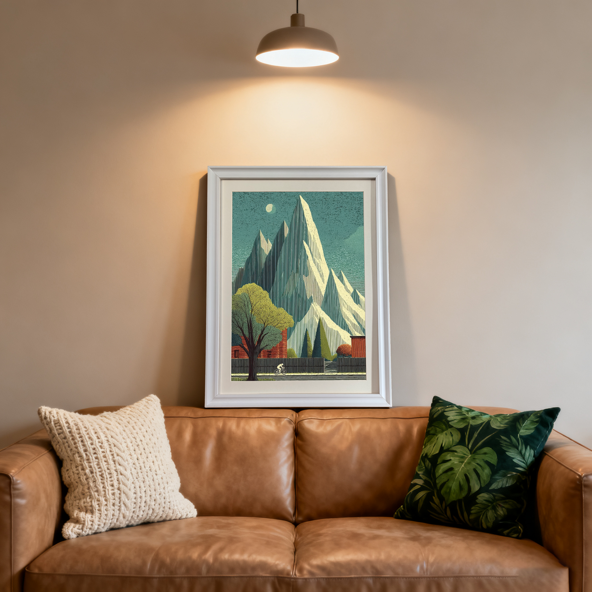 White-framed art print of a striking mountain landscape with a lone cyclist, a large tree, and red buildings. It rests on a brown leather couch in a modern room, illuminated by a spotlight.
