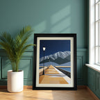 Geometric art print of a pier leading into the distance toward mountains, in a thick black frame, leaning on a wooden floor against a dark teal paneled wall next to a potted palm plant.
