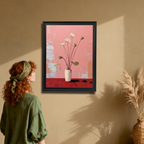 A young woman with red, curly hair and a green headband stands in an earth-toned room looking up at a vertical, black-framed painting of white lotus flowers on a pink and red background hanging on the wall. A woven vase with dried grasses is on the right.