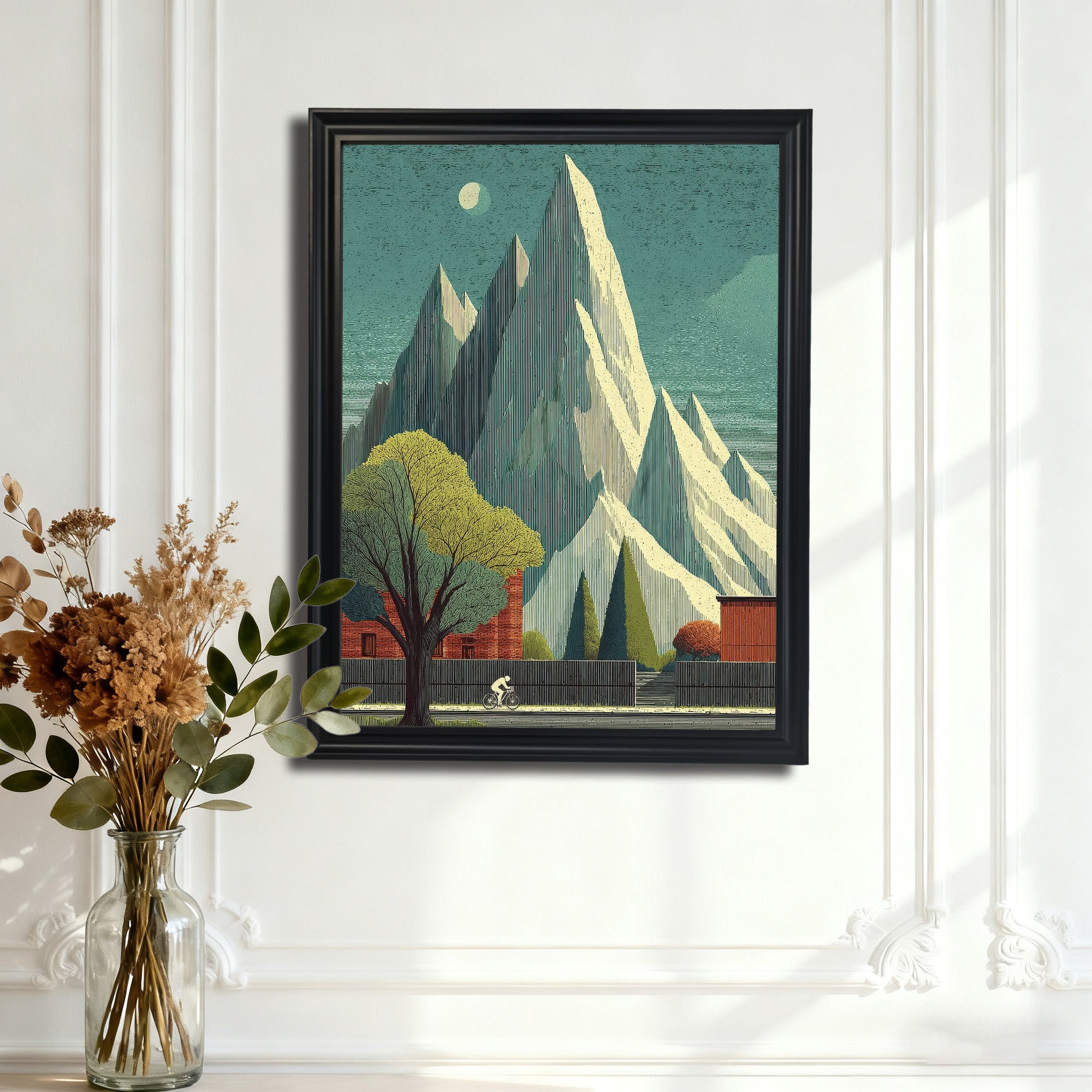 Black-framed art print of the tall, jagged mountain landscape with a cyclist, hanging on an ornate white paneled wall above a surface with a clear vase of dried botanicals.