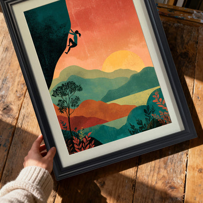 Close-up shot of hands holding a framed art print of a climber silhouette against a sunset landscape in a thick black wood frame, over a rustic wooden table.