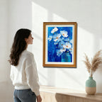 A woman in a white sweater stands in a bright room looking at a gold-framed painting of white flowers in a blue jar against a bright blue background, mounted above a light wood console table.
