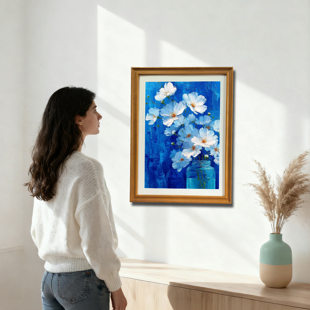 A woman in a white sweater stands in a bright room looking at a gold-framed painting of white flowers in a blue jar against a bright blue background, mounted above a light wood console table.