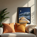 Geometric style art print of a long perspective pier with glowing streetlights leading toward sharp, snow-capped mountains under a dark blue sky, framed in black, resting on a white sofa with orange pillows.
