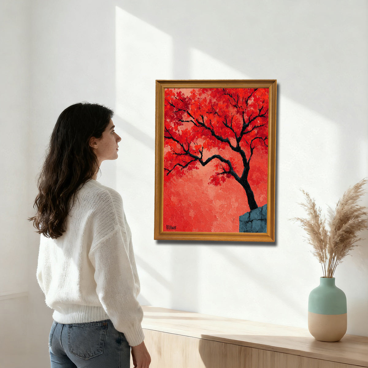 A woman in a white sweater stands in a bright, modern room looking at a gold-framed painting of a textured red-orange tree mounted above a light wood console table.