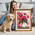 A smiling woman sits on a shag rug next to a golden retriever puppy, holding a gold-framed painting of pink roses in a vase against a distressed pink/white background. A gray sofa is visible behind them.