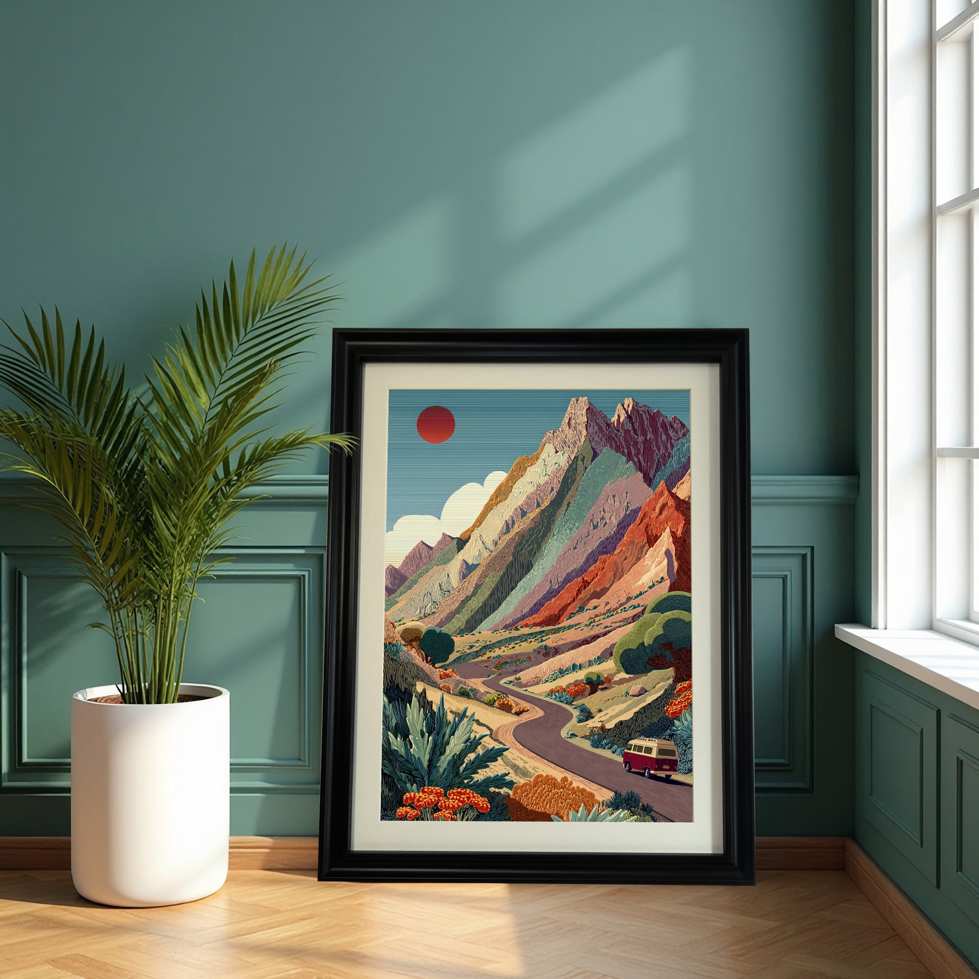 Large, black-framed art print of the colorful mountain road trip scene resting on the floor next to a potted palm, against a teal green paneled wall.