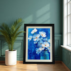 A large, black-framed painting of white flowers in a blue jar against a vivid blue background is leaning on a hardwood floor against a dark teal green paneled wall, beside a large potted palm plant.