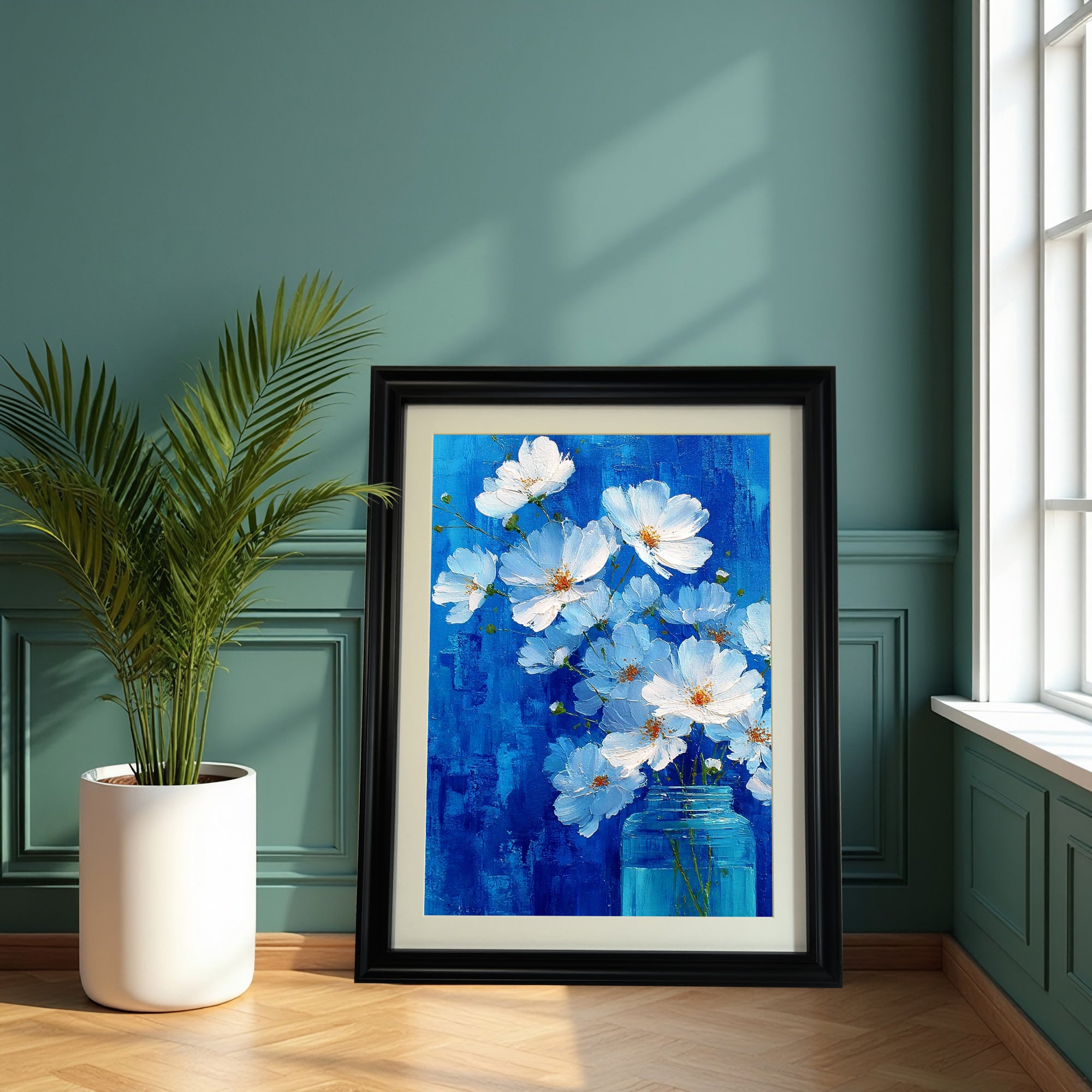 A large, black-framed painting of white flowers in a blue jar against a vivid blue background is leaning on a hardwood floor against a dark teal green paneled wall, beside a large potted palm plant.