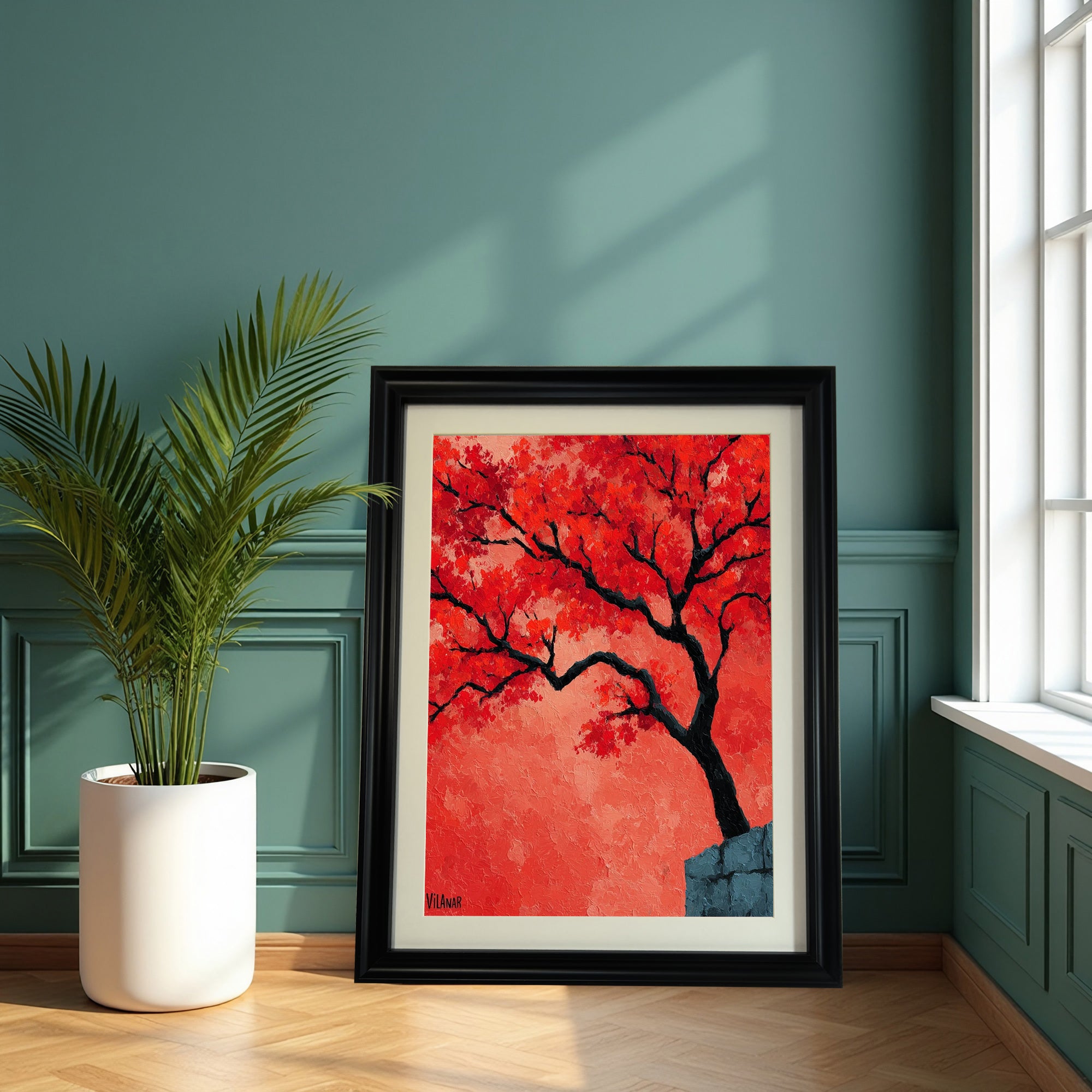 A large, black-framed painting of a vibrant red-orange tree is leaning on a hardwood floor against a dark teal green paneled wall, beside a large potted palm plant in a white container.