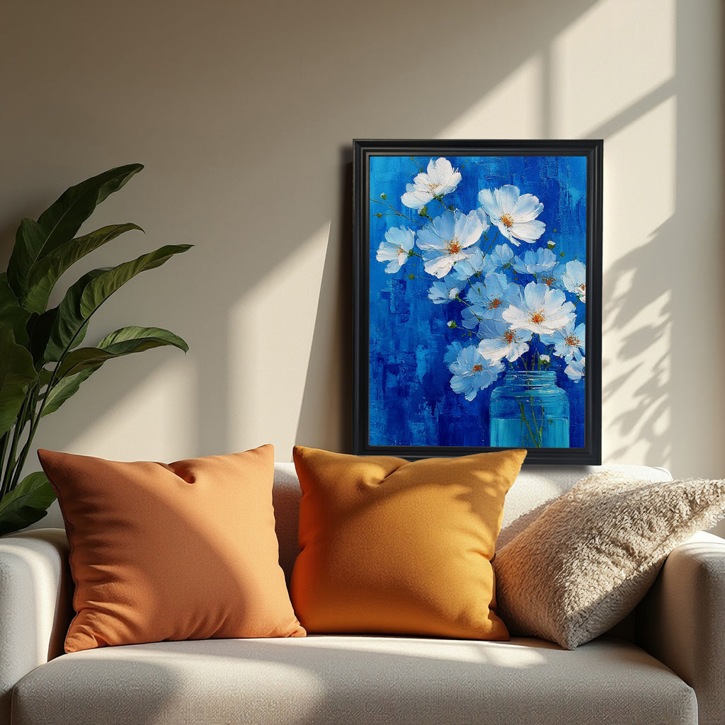 A black-framed painting of white flowers in a blue jar against a vivid blue background is resting on a beige sofa, flanked by two orange/mustard pillows. A large green houseplant is visible on the left.