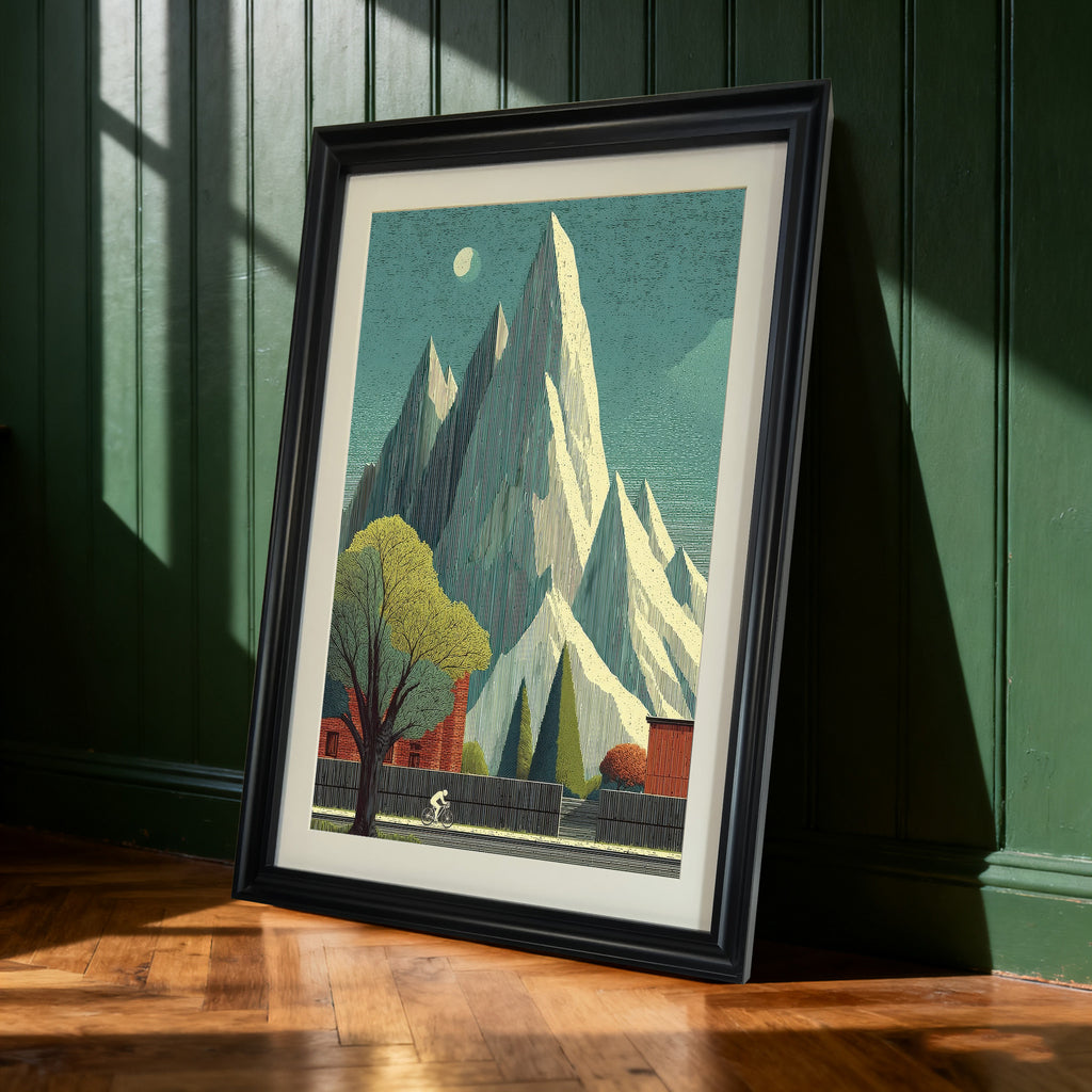 Black-framed art print of the tall mountain and cyclist landscape leaning against a dark green paneled wall. Sunlight casts shadows on the wall and wooden floor.