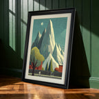 Black-framed art print of the tall mountain and cyclist landscape leaning against a dark green paneled wall. Sunlight casts shadows on the wall and wooden floor.