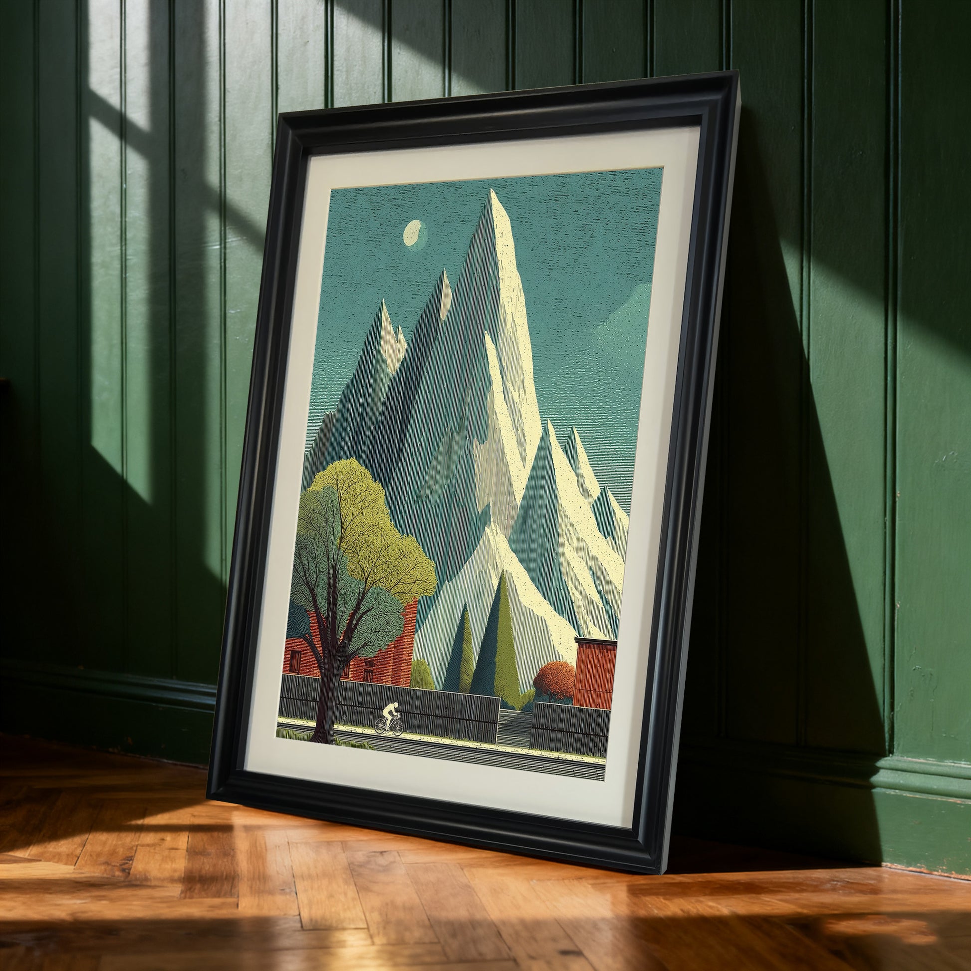 Black-framed art print of the tall mountain and cyclist landscape leaning against a dark green paneled wall. Sunlight casts shadows on the wall and wooden floor.