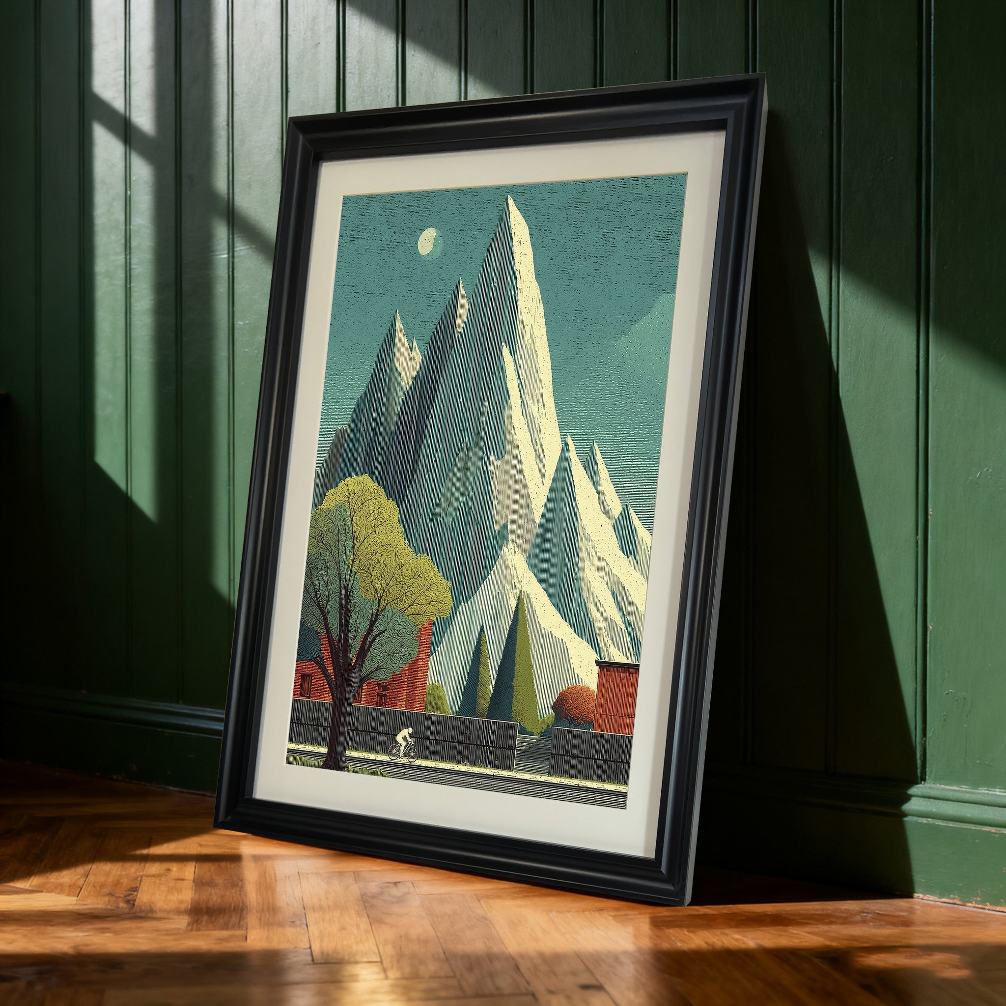 Black-framed art print of the tall mountain and cyclist landscape leaning against a dark green paneled wall. Sunlight casts shadows on the wall and wooden floor.