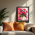 A black-framed painting of pink roses in a vase against a distressed pink/white background is resting on a beige sofa, framed by two orange/yellow pillows. A large green houseplant is visible on the left.
