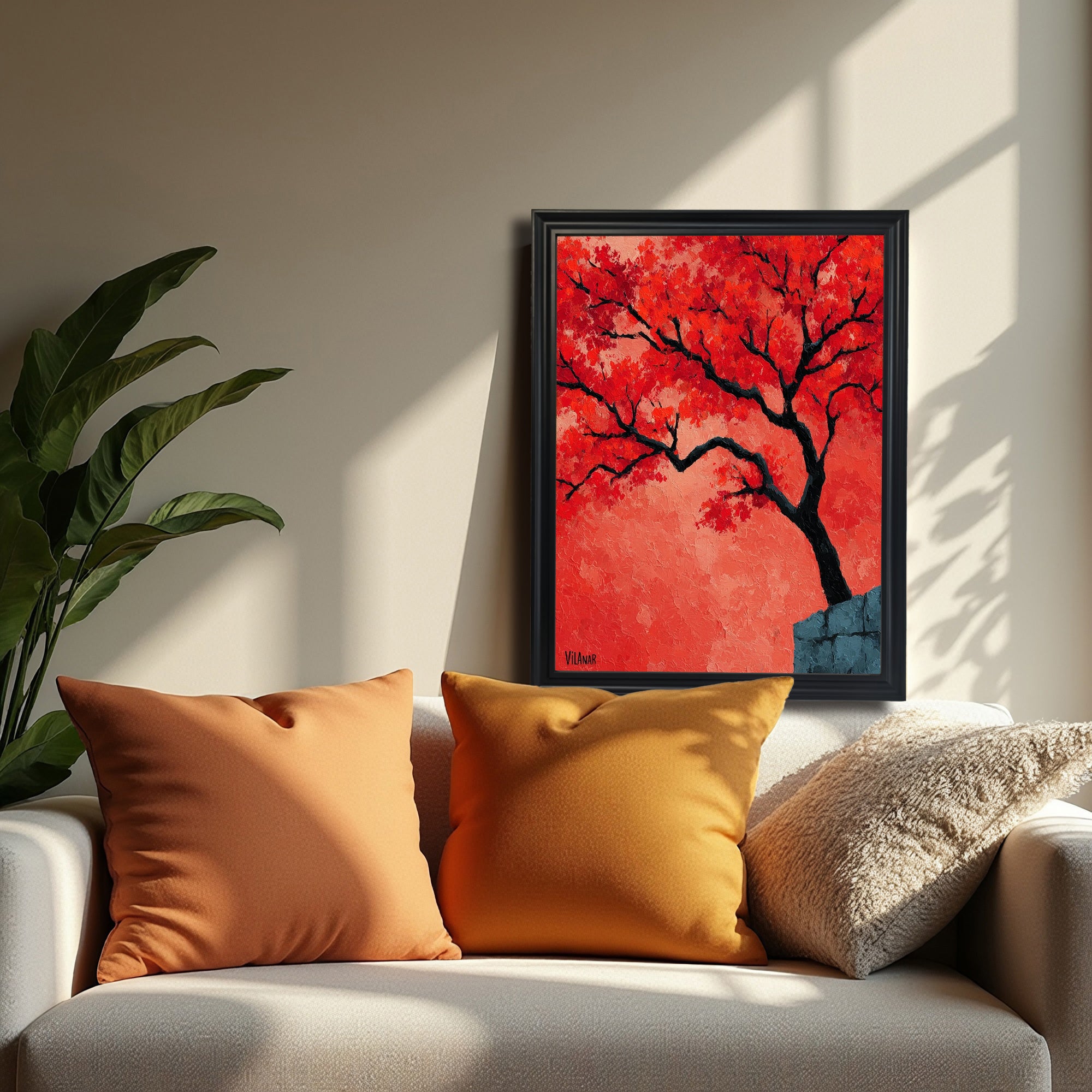 A black-framed painting of a vibrant red-orange tree is resting on a beige sofa, framed by two mustard yellow pillows. A large green houseplant is visible on the left.