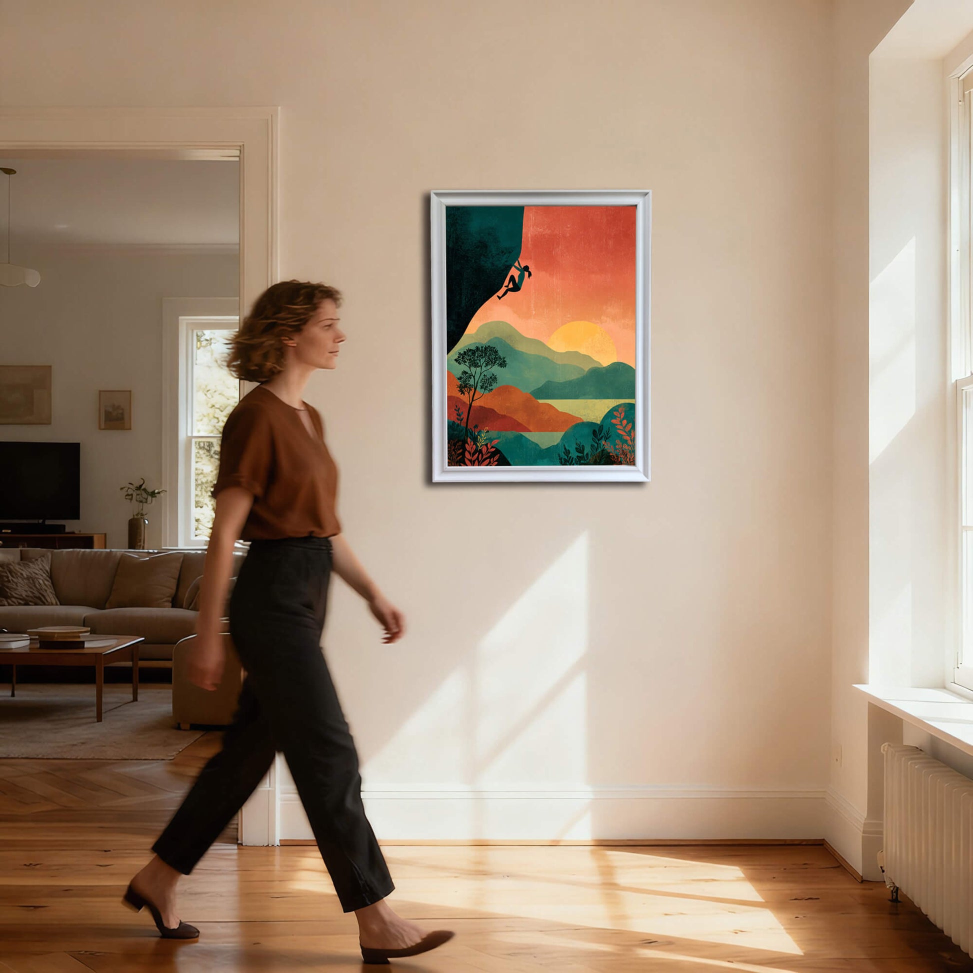 Full-body shot of a woman walking past a vertical art print of a climber silhouette and sunset landscape, framed in a thin white frame, hanging on a cream wall in a sunlit hallway.
