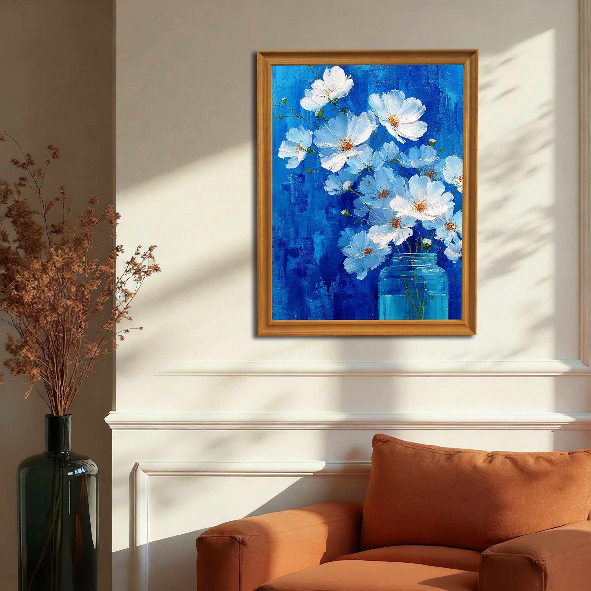 A vertical, gold-framed painting of white flowers in a blue jar against a vibrant blue background is hanging on a beige wall with white wainscoting, positioned above an orange chair