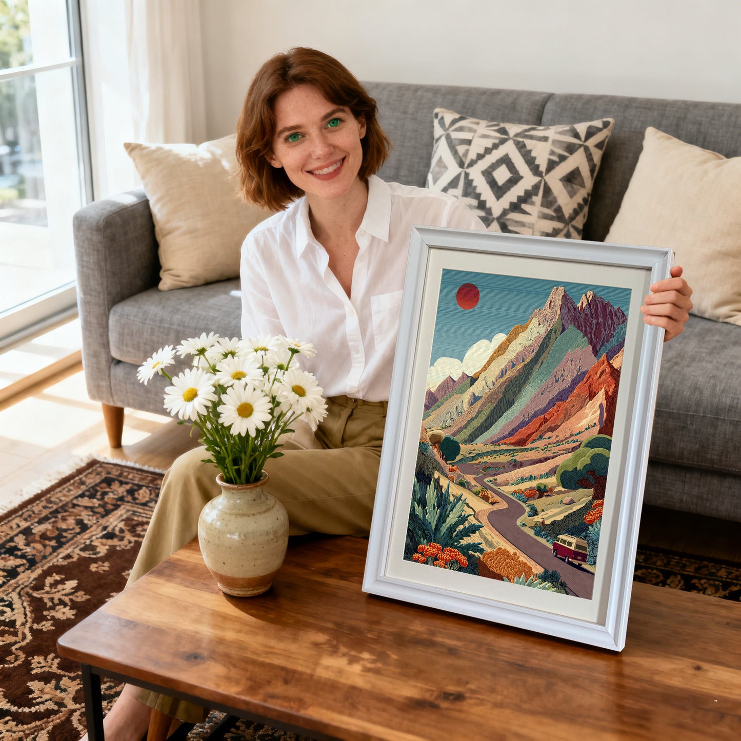 Mountain Road Trip,"Smiling woman sitting on a couch, holding a white-framed art print of the colorful mountain landscape with a winding road and a red van. Daisies are on the coffee table.