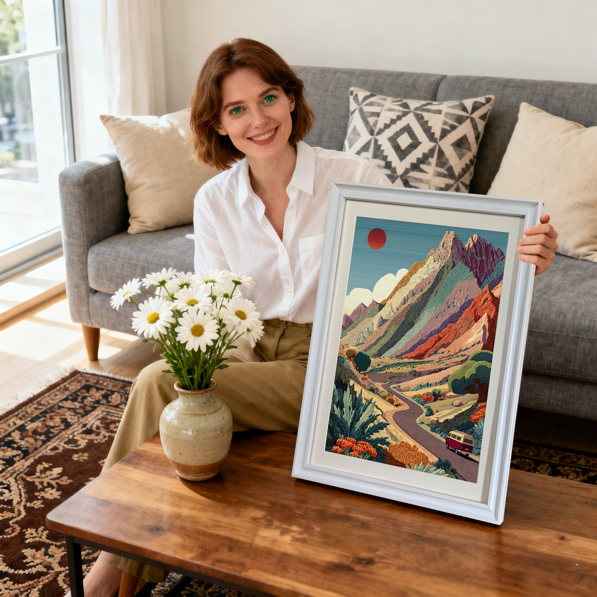 Mountain Road Trip,"Smiling woman sitting on a couch, holding a white-framed art print of the colorful mountain landscape with a winding road and a red van. Daisies are on the coffee table.