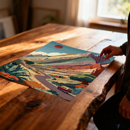 Unframed art print showing a colorful, layered mountain landscape with a road and van, resting on a rustic wooden table. A person's hand is visible near the edge.