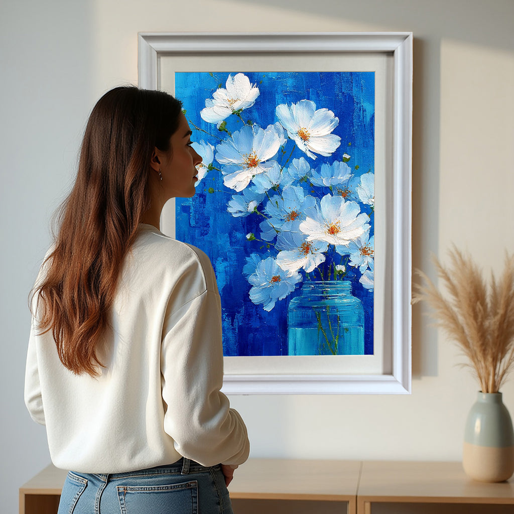 A woman in a white sweater stands looking at a large, white-framed painting of white flowers in a blue jar against a textured blue background, hanging above a light wood console table in a bright room.